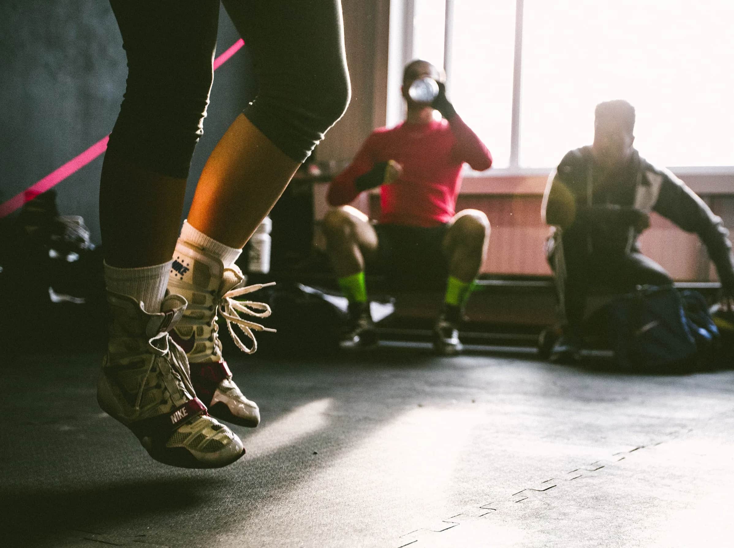 Close-up of a person jumping rope in a gym, wearing boxing shoes, while two people sit in the background near large windows, one drinking from a bottle. Sunlight shines through the window, casting shadows on the floor.