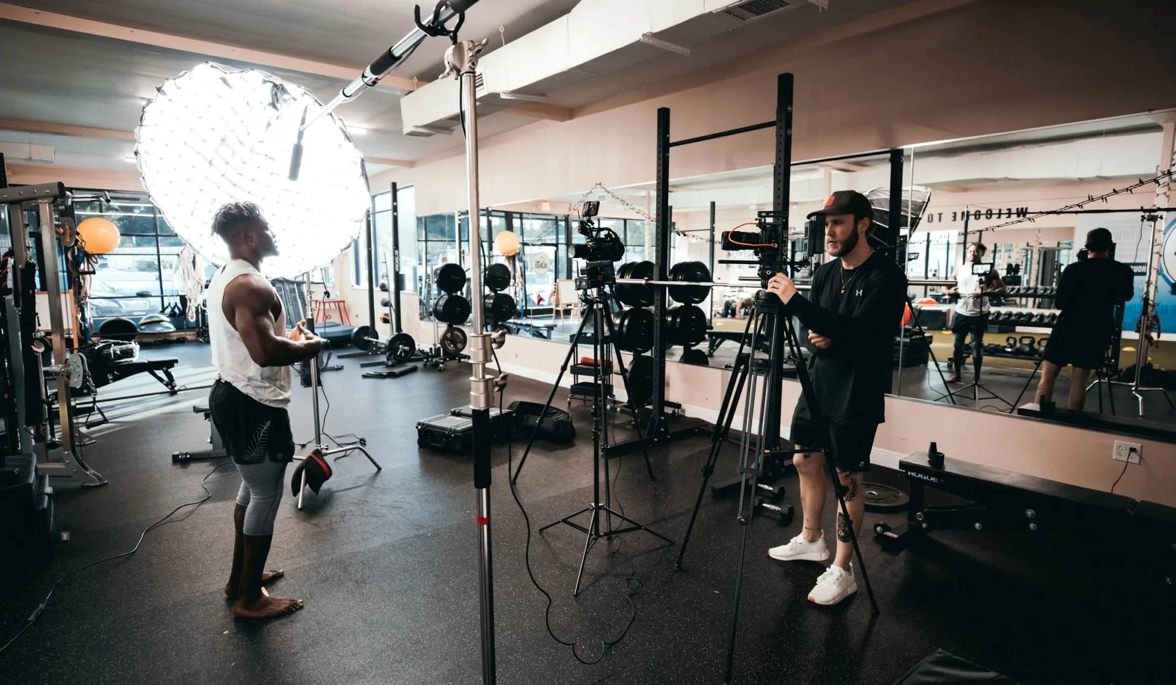 A person in athletic wear stands in a gym, being filmed by another person with a camera on a tripod. Large lights and gym equipment are visible around them, and mirrors line the walls.