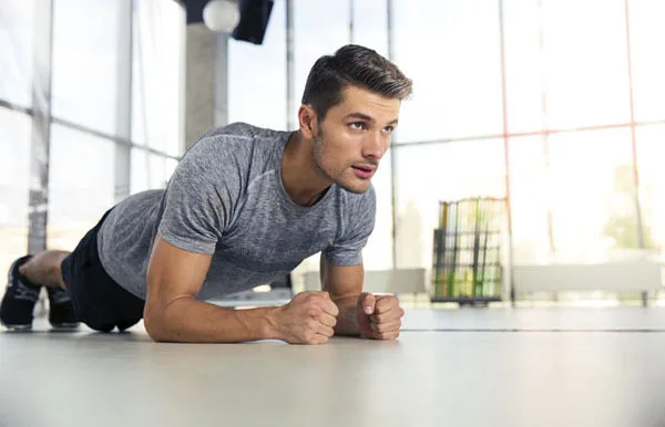 A man in a gray t-shirt and black shorts is holding a plank position on the floor in a bright, spacious gym with large windows.