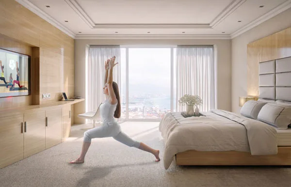 A woman practices yoga in a bright, modern bedroom, performing a lunge pose while facing a TV displaying a yoga video. Large windows behind her reveal a scenic outdoor view.