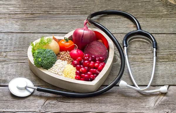 A heart-shaped wooden bowl filled with colorful vegetables, grains, and berries sits on a rustic wooden surface, next to a stethoscope, symbolizing healthy eating and heart health.