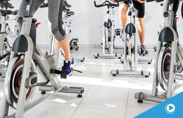 People exercising on stationary bikes in a bright, clean indoor cycling studio. The focus is on their legs and the bikes as they pedal.