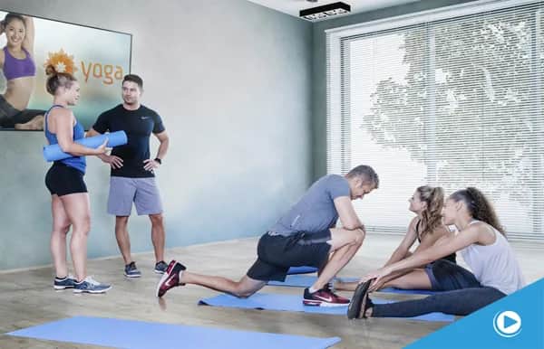 Four people in athletic wear are stretching and talking on yoga mats in a bright studio with large windows; a yoga poster is on the wall behind them.
