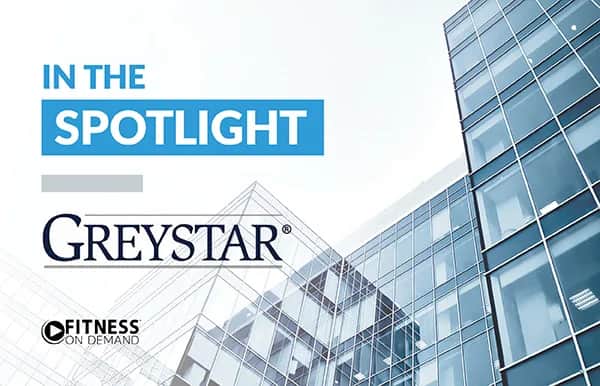 Modern glass office building with text: IN THE SPOTLIGHT, Greystar, and Fitness On Demand logo on the left side against a light blue sky.