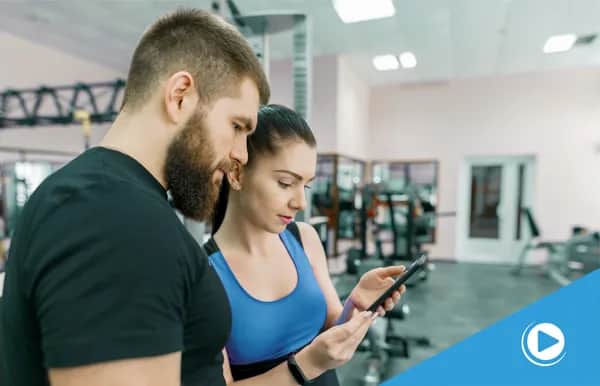 A man and a woman in athletic wear stand together in a gym, looking at a smartphone. Exercise equipment and weights are visible in the background. A blue corner with a play icon is in the bottom right.