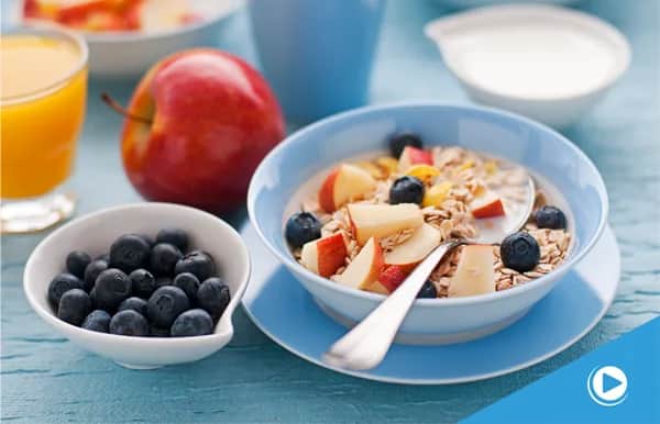 A bowl of muesli with milk, apple slices, and blueberries on a blue plate, next to a bowl of fresh blueberries, a red apple, a glass of orange juice, and a small bowl of yogurt on a blue tablecloth.