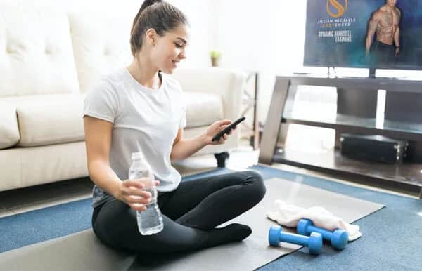 A woman in workout clothes sits cross-legged on a yoga mat, holding a water bottle and looking at her phone. Two blue dumbbells and a towel are on the mat, and a fitness video plays on a TV in the background.