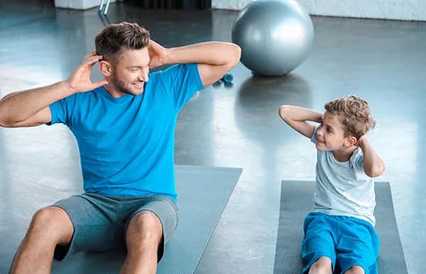 A man and a young boy are sitting on exercise mats indoors, doing sit-ups together. Both are smiling and looking at each other, with their hands behind their heads. An exercise ball and dumbbells are in the background.