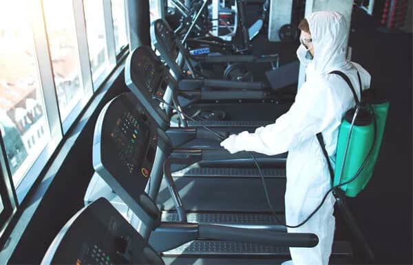 A person in full protective gear disinfects treadmills at a gym, spraying cleaner from a green backpack sprayer, with sunlight coming through large windows.