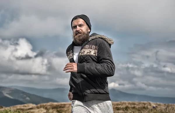 A bearded man wearing a knit hat, patterned jacket, and sweatpants is jogging outdoors with cloudy skies and mountains in the background.