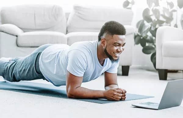A smiling person in workout clothes holds a plank position on a yoga mat indoors, looking at a laptop screen in front of them. A sofa and a plant are visible in the background.