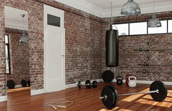 A home gym with brick walls, wooden floor, and large windows. Equipment includes a barbell, dumbbells, kettlebell, punching bag, jump rope, and weights on racks. A mirror and white door are also visible.
