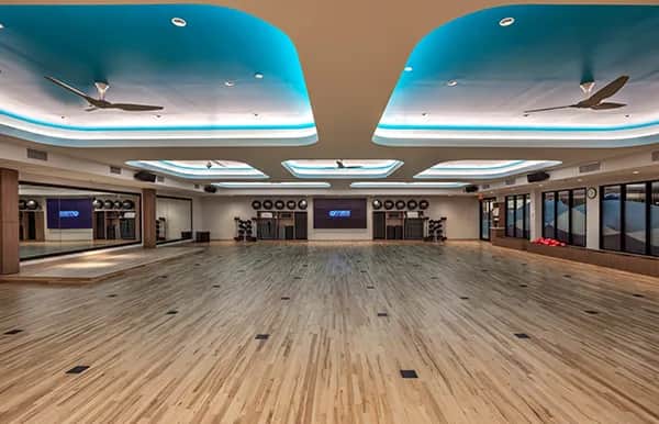 Spacious fitness studio with a polished wooden floor, mirrored walls, ceiling fans, and blue accent lighting. Exercise equipment and two large TVs are visible at the far end of the room.