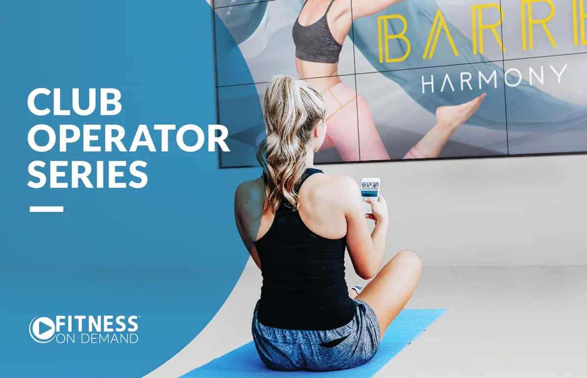 A woman sits on a blue yoga mat in a gym, holding a phone and facing a large screen displaying a barre fitness class. Text on the left reads Club Operator Series and Fitness On Demand.