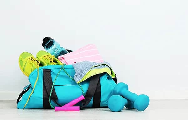 A turquoise gym bag with green shoes, a water bottle, a pink towel, gray clothing, pink hand grips, and a pair of blue dumbbells arranged on the floor against a white background.