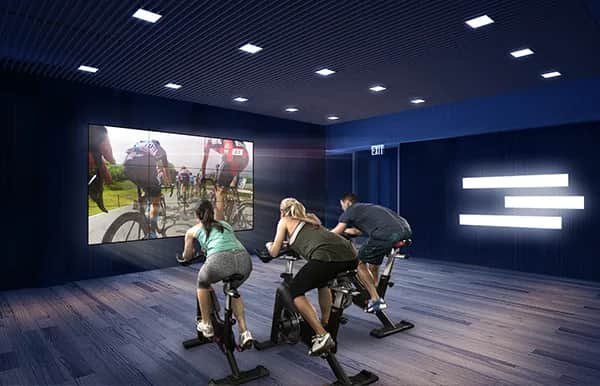 Three people ride stationary bikes in a dark, modern fitness room, facing a large screen displaying a virtual cycling scene. Blue walls and ceiling lights create an immersive atmosphere.