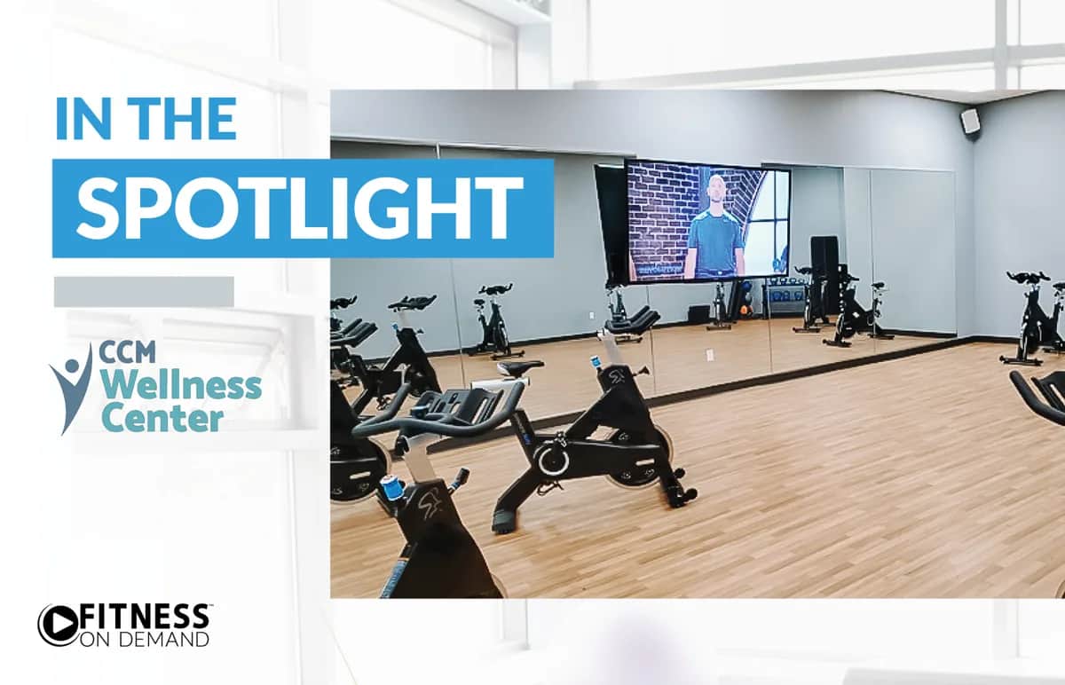 A modern fitness studio with wooden floors, several stationary bikes, and a large mirror. A screen shows a virtual instructor. Text reads IN THE SPOTLIGHT, CCM Wellness Center, and Fitness On Demand.