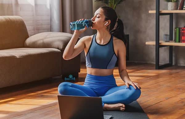 A woman in workout clothes sits cross-legged on a yoga mat indoors, drinking water from a bottle and wearing wireless earbuds, with an open laptop in front of her. Sunlight streams through a window behind her.