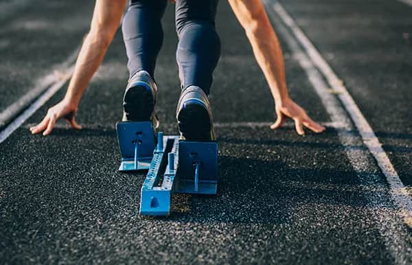 Close-up of a sprinter at the starting blocks on a track, shown from behind, with hands and feet positioned ready to launch at the beginning of a race.