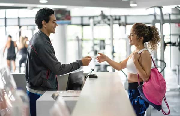 A woman in workout clothes hands her gym card to a smiling receptionist at a gym entrance, with exercise equipment and people in the background.