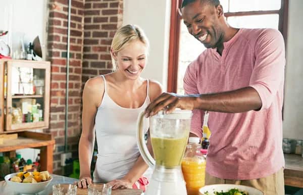 A smiling man and woman stand in a kitchen, blending a green smoothie in a blender. The kitchen has brick walls and a window, with fruit and glasses on the counter.