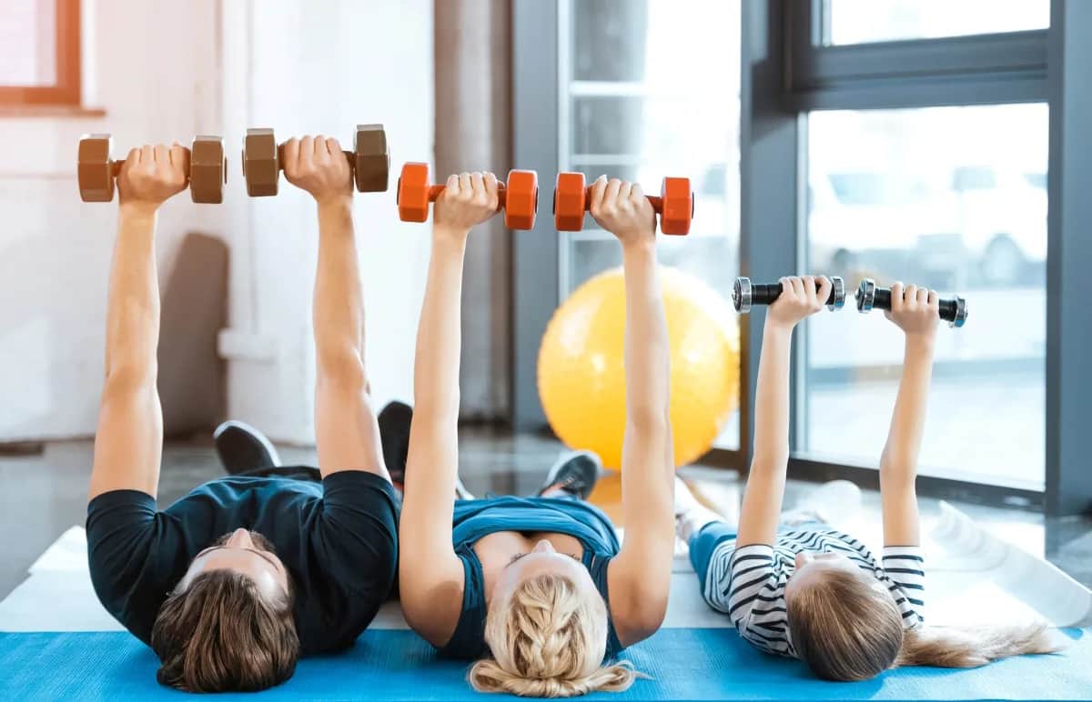 Three people, two adults and a child, lie on their backs on yoga mats indoors, lifting dumbbells above their chests. A large yellow exercise ball sits in the background near large windows.