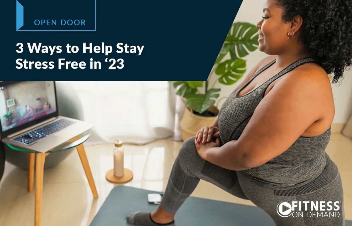 A woman in workout clothes sits on a yoga mat at home, watching a fitness video on her laptop. The text overlay reads, 3 Ways to Help Stay Stress Free in ‘23 and Fitness On Demand.