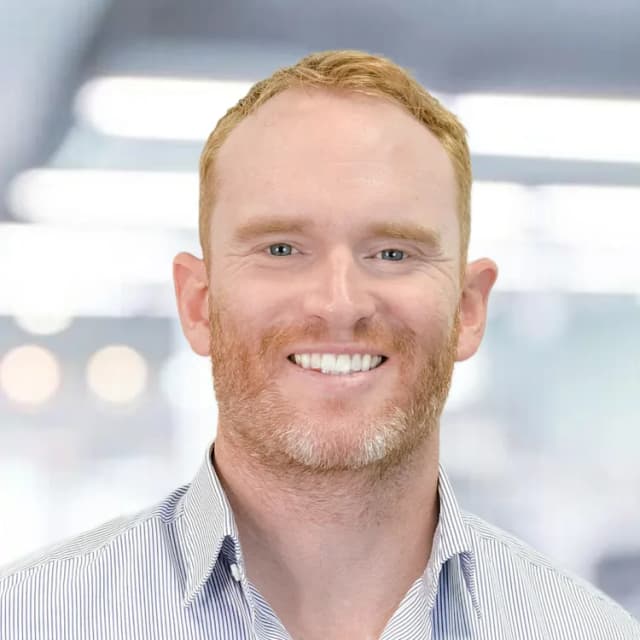 A smiling man with short red hair and a trimmed beard, wearing a light blue and white striped collared shirt, stands in front of a blurry, brightly lit indoor background.