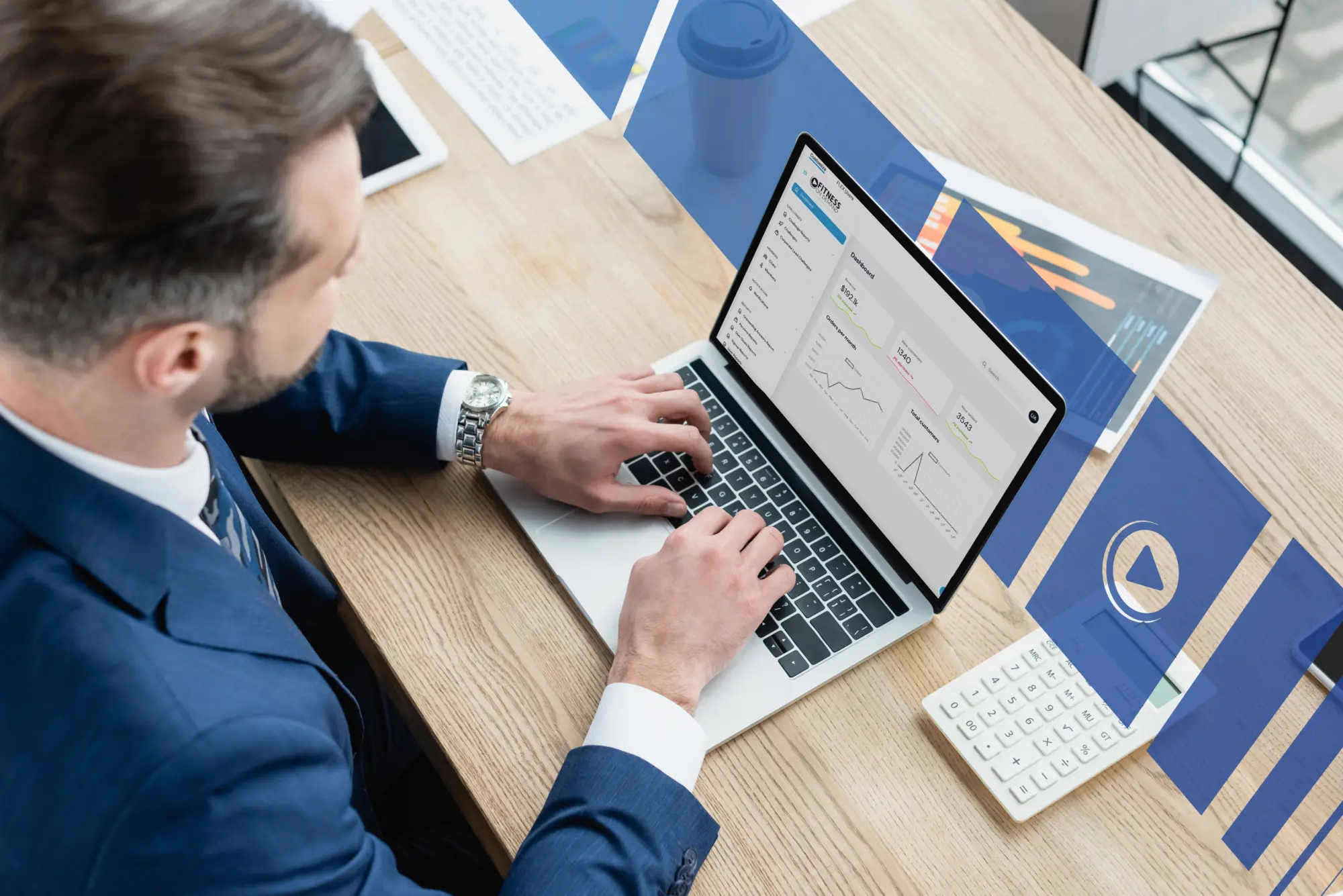 A man in a suit works on a laptop at a wooden desk, analyzing graphs and charts on the screen. The desk has papers, a calculator, a smartphone, and a blue coffee cup.