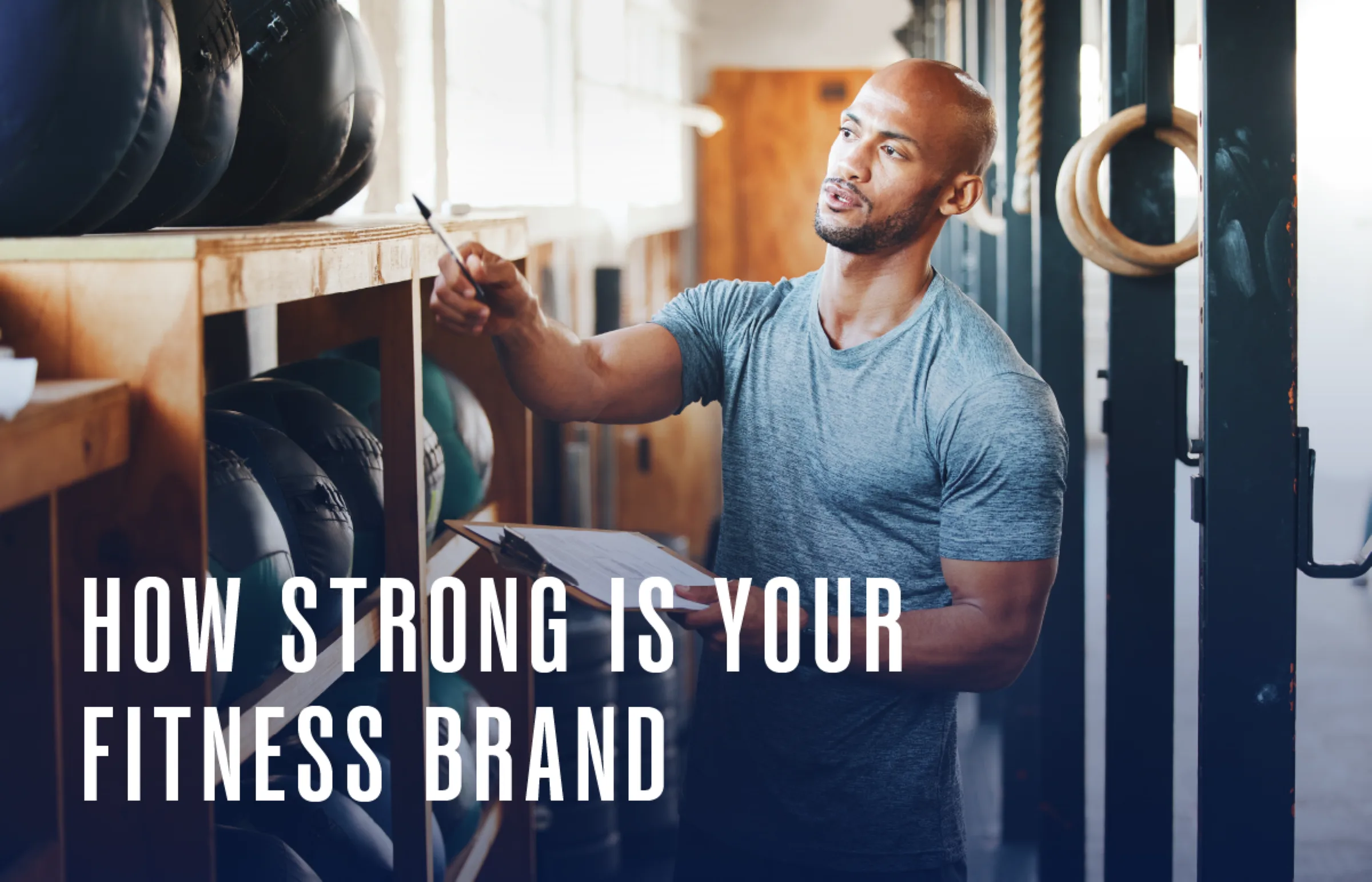 A man in a gym wearing a gray t-shirt holds a clipboard and pen while checking medicine balls on a shelf. Text on the image reads, How strong is your fitness brand. Gym equipment hangs in the background.