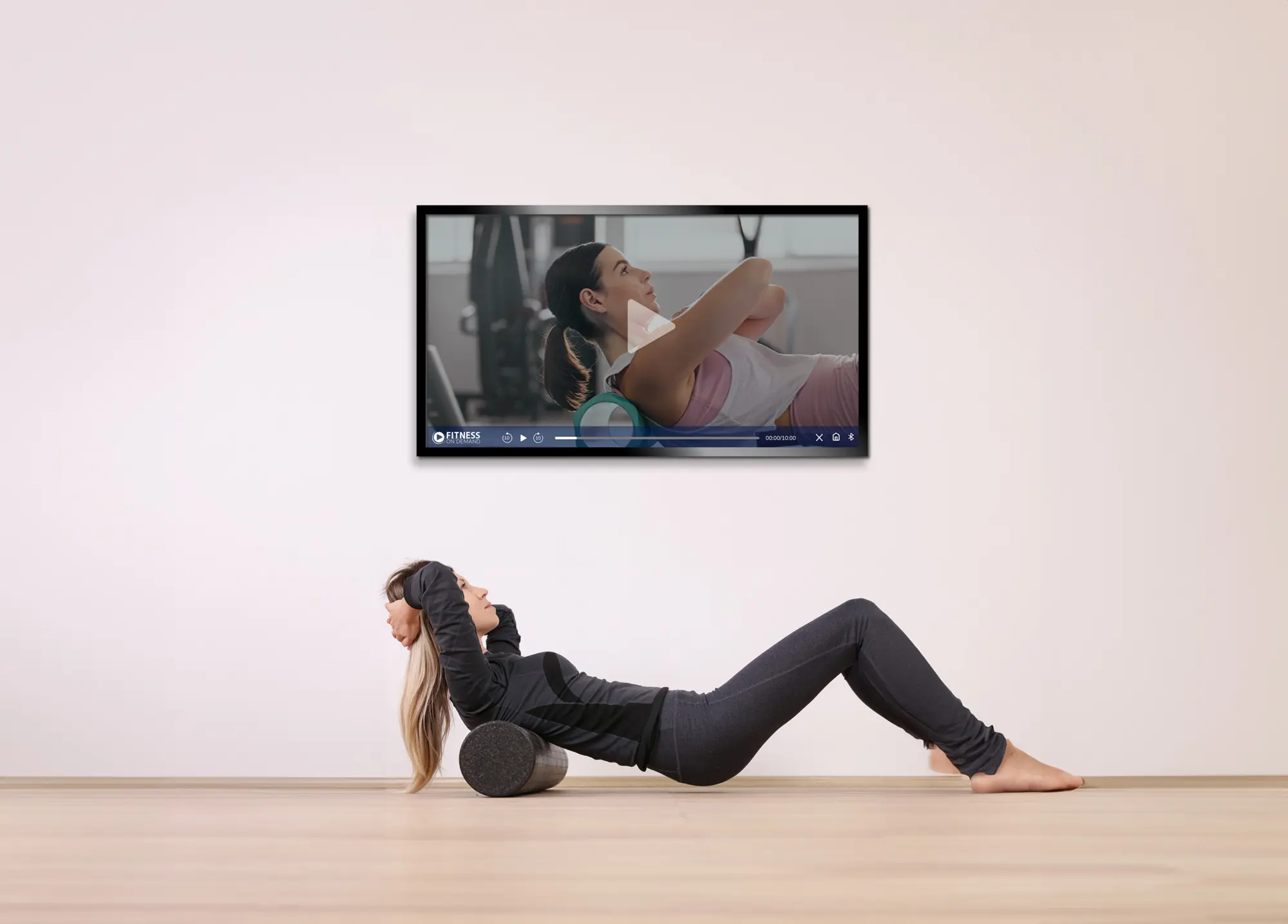 A woman lying on a foam roller in a minimal room follows an exercise video displayed on a wall-mounted TV, mimicking the workout shown on the screen.
