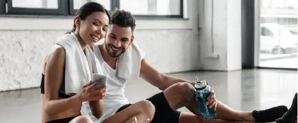 A smiling woman and man in workout clothes sit on the gym floor with towels around their necks. The woman holds a phone, and the man holds a water bottle. They appear to be looking at something on the phone together.