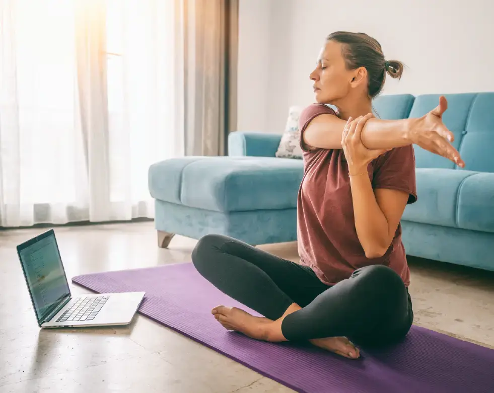 A woman sits cross-legged on a yoga mat in her living room, stretching her arm across her chest while following an online workout on her laptop.