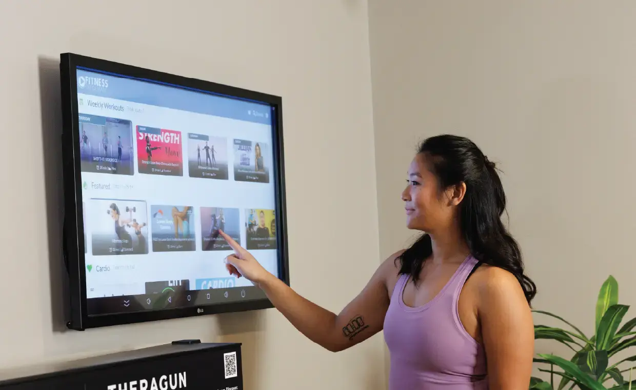 A woman in a purple tank top touches a wall-mounted touchscreen displaying various workout videos, selecting an option.
