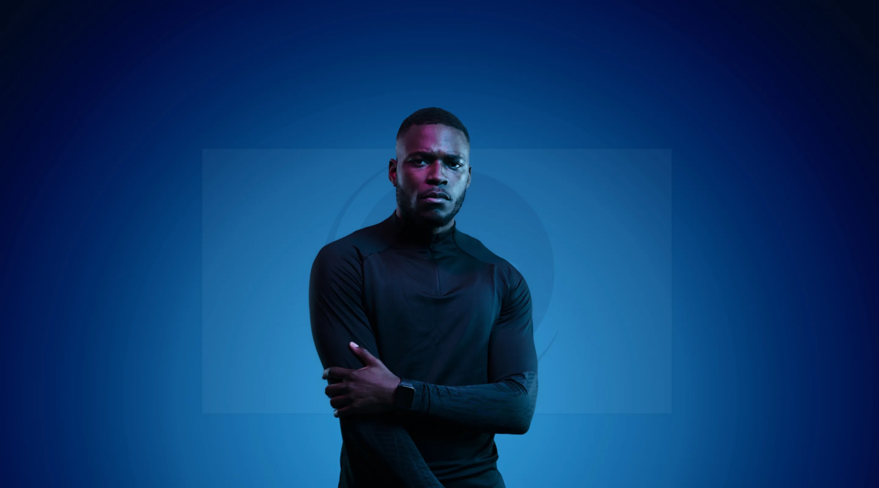 A man wearing a black long-sleeve athletic shirt stands against a blue gradient background, looking forward with a serious expression. One arm is crossed over his body, and he wears a smart watch on his wrist.