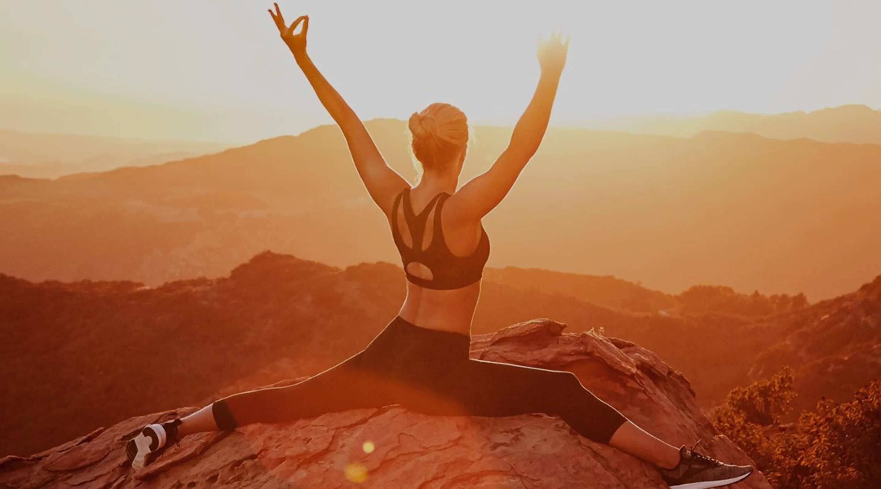 A person in athletic wear does a split on a rocky mountaintop at sunrise, arms raised in celebration, overlooking scenic hills and valleys bathed in golden light.