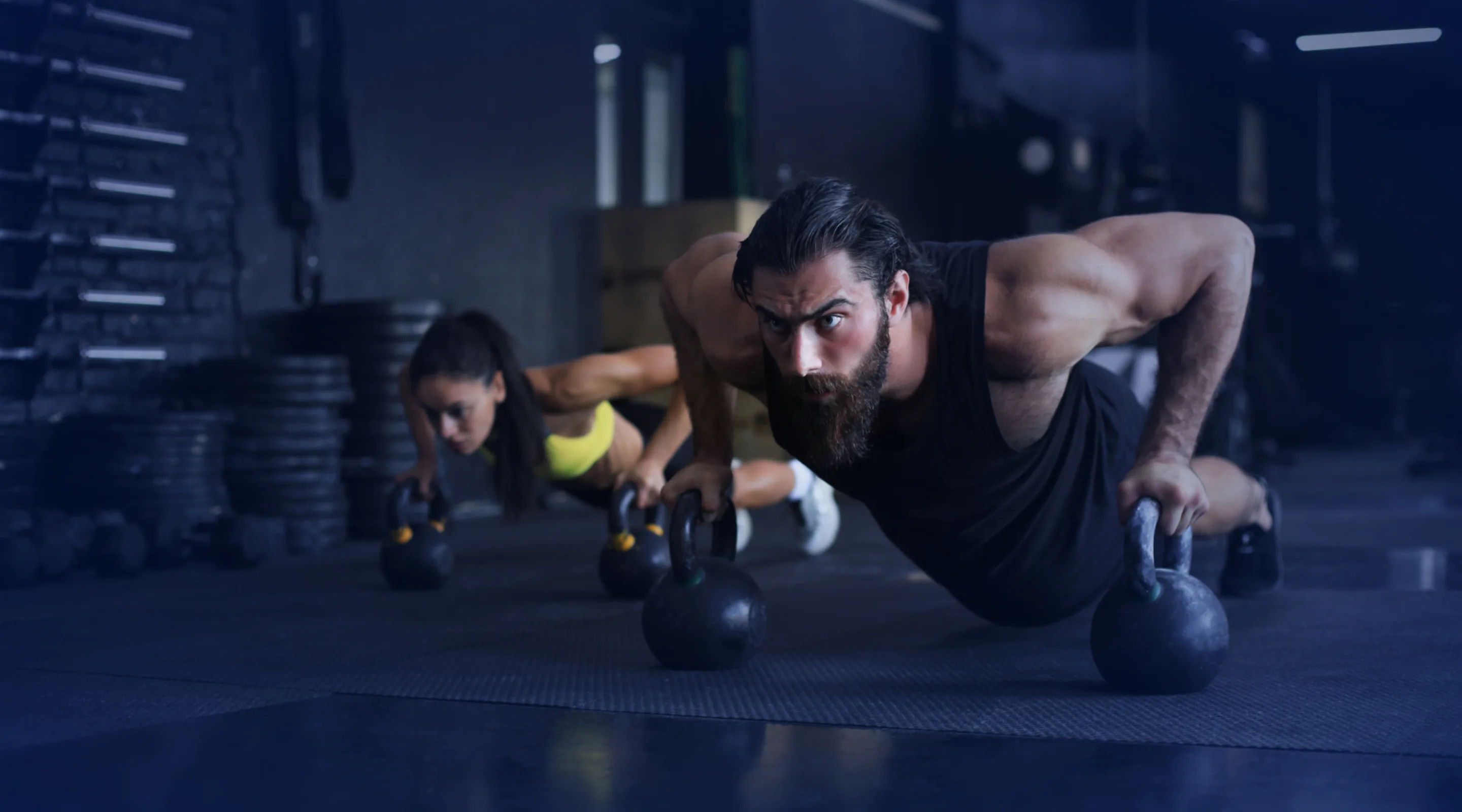 A man and a woman exercising in a gym, performing push-ups while gripping kettlebells, with focused expressions and workout equipment visible in the background.