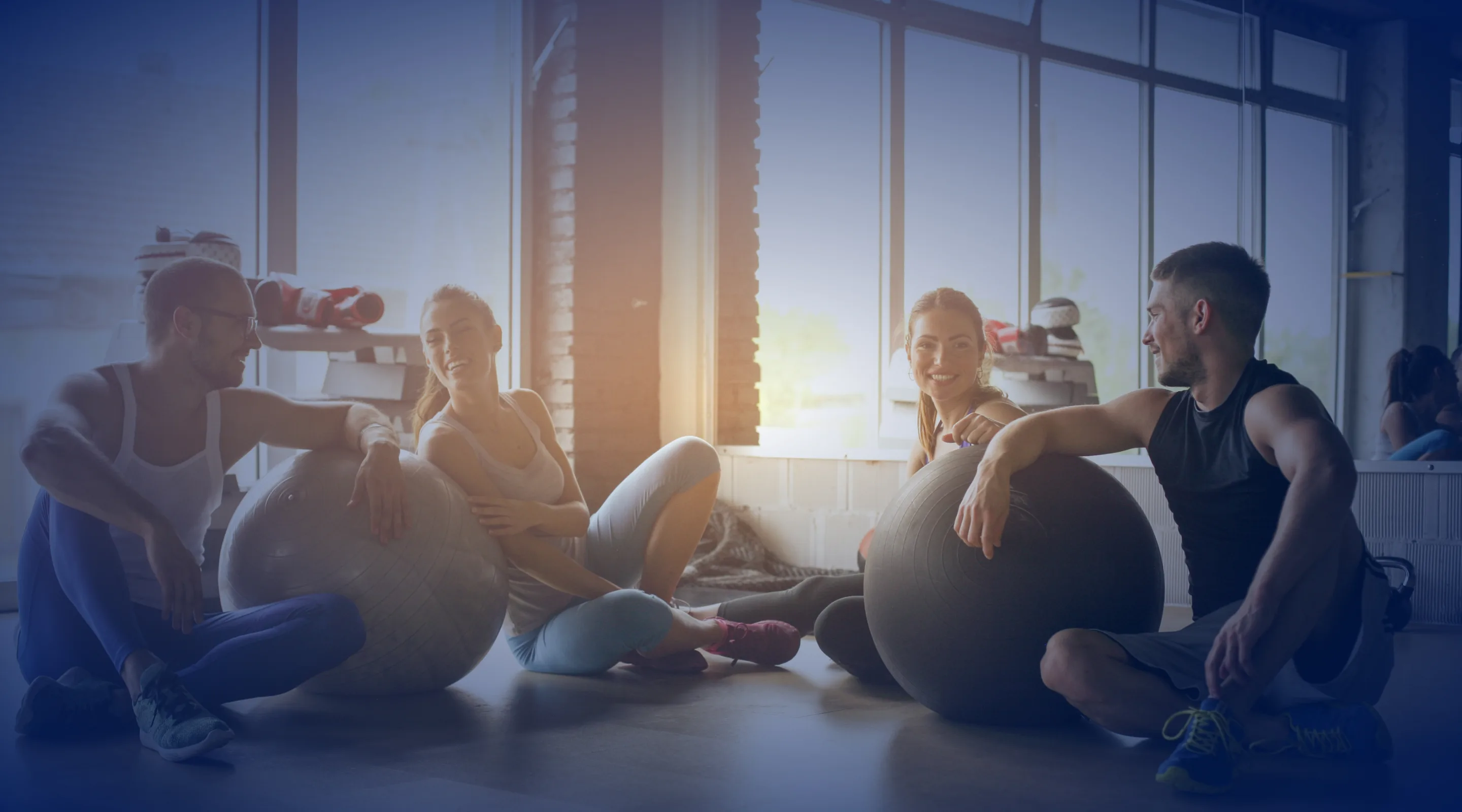 Four people in workout clothes sit on the floor of a gym, smiling and talking, with large exercise balls in front of them and sunlight streaming through big windows in the background.