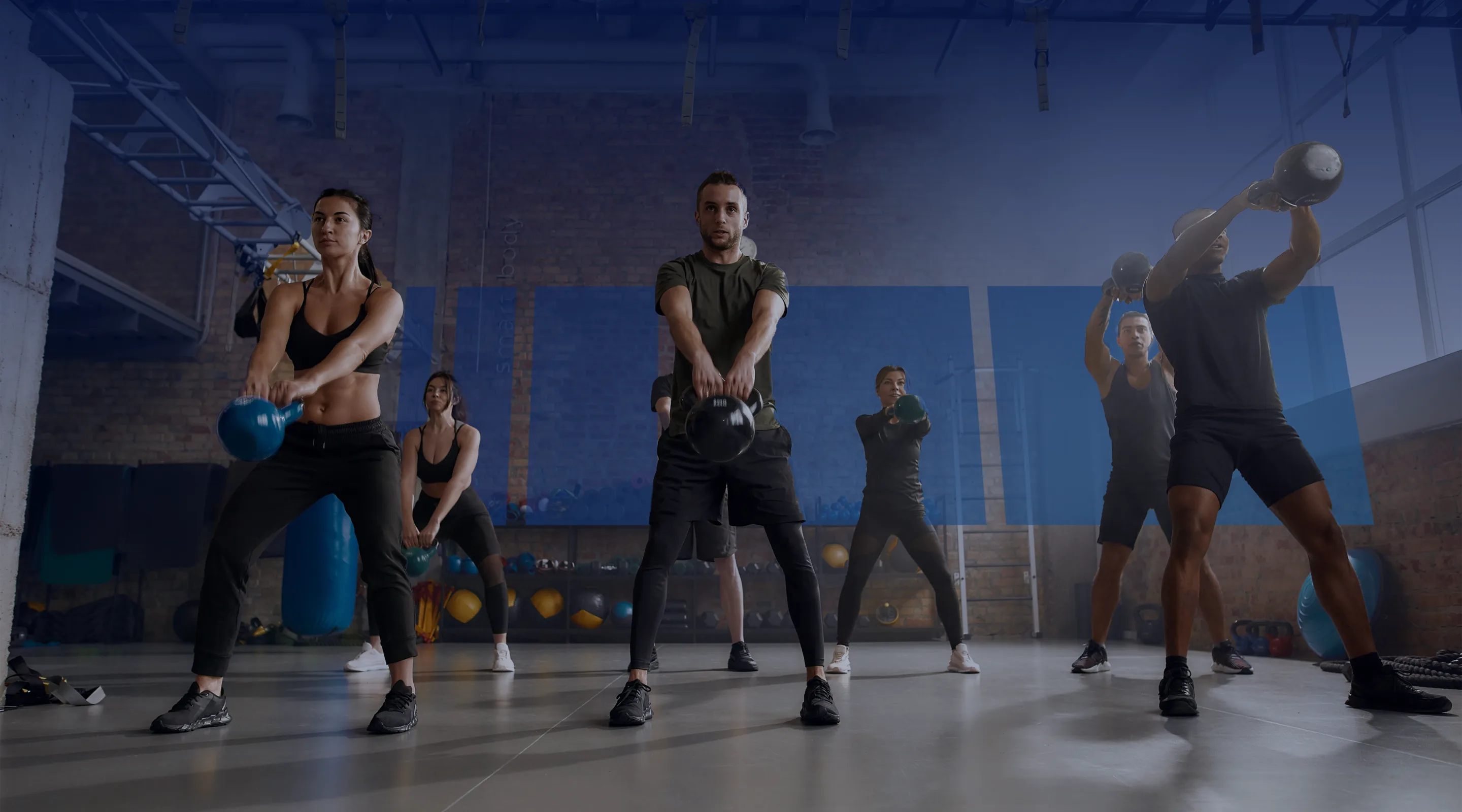 A group of people in athletic wear exercise together indoors, each lifting kettlebells in unison during a fitness class. The gym has brick walls and workout equipment in the background.