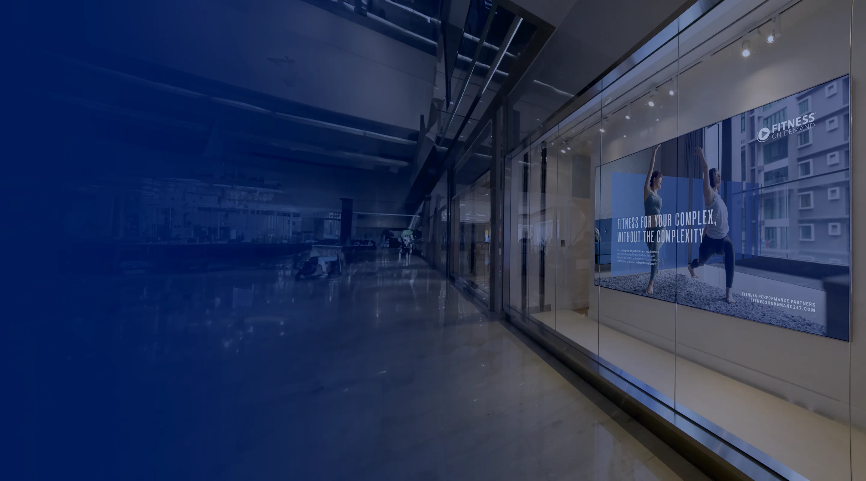 A modern shopping mall hallway with a brightly lit fitness advertisement on a glass wall, showing two people exercising and the text Stronger mind. Complex, intuitive capability.