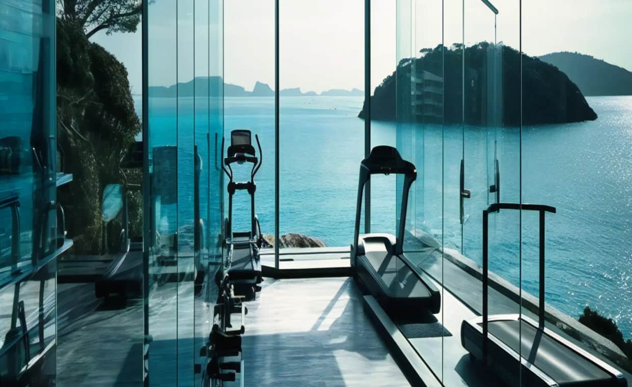 A modern glass-walled gym with treadmills and elliptical machines overlooks a scenic ocean view with islands and blue water under a clear sky.