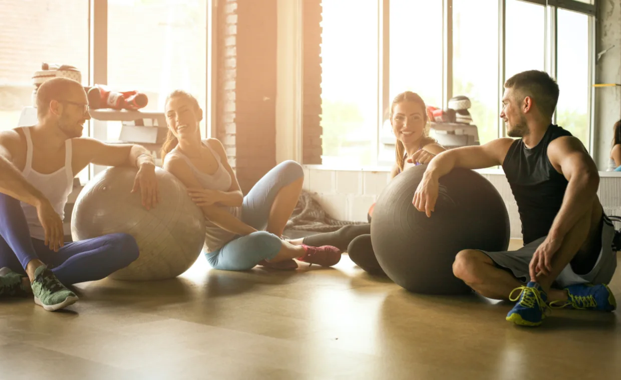 Four people sit on a gym floor, smiling and talking while leaning on large exercise balls, with sunlight streaming through windows in the background.
