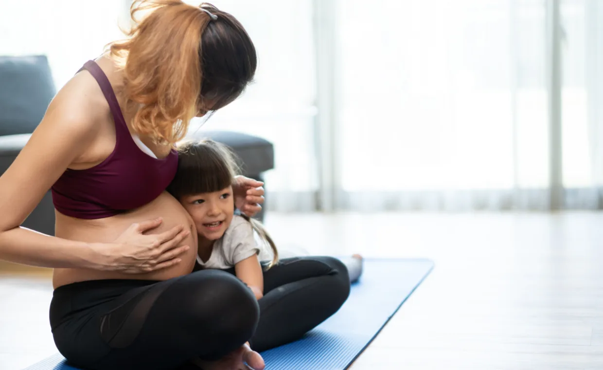 A young child hugs and rests her head on the pregnant belly of a woman sitting on a yoga mat. Both appear happy in a bright, softly lit room.