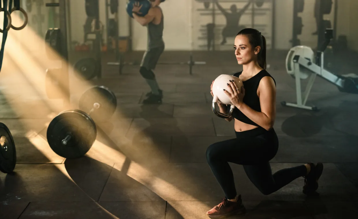 A woman in athletic wear performs a lunge while holding a medicine ball in a gym. Sunlight streams through the windows. Gym equipment and another person working out are visible in the background.
