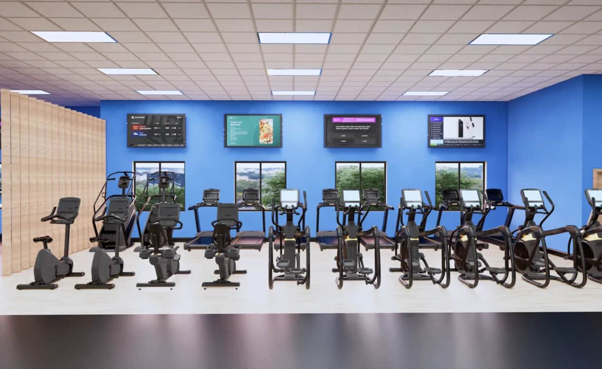 A row of stationary exercise bikes and elliptical machines is lined up in a brightly lit gym with blue walls, large windows, and TV screens mounted above the equipment.
