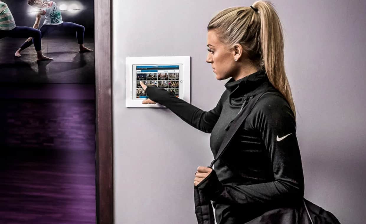 A woman with a ponytail and a gym bag uses a touchscreen panel on a wall, selecting from exercise or class options. In the background, people are doing yoga or stretching in a dimly lit studio.