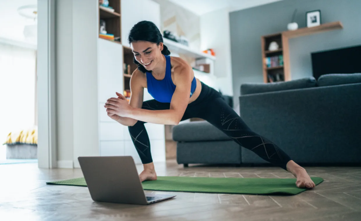 A woman in athletic wear stretches on a yoga mat in her living room while following an exercise video on a laptop. She smiles and leans to one side with hands clasped, with shelves and a couch in the background.