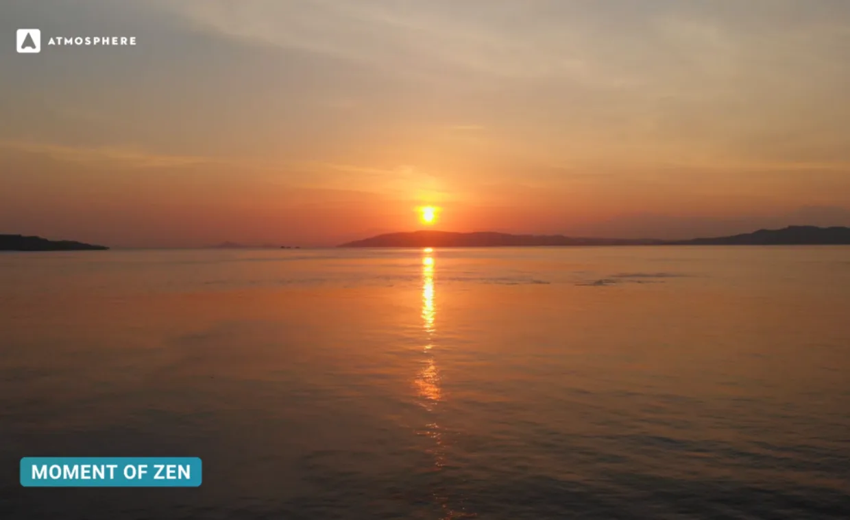 A calm sunset over the ocean, with the sun low above distant hills, casting a golden reflection on the water. The sky is painted with warm orange hues. Text: ATMOSPHERE in the top left and MOMENT OF ZEN in the bottom left.