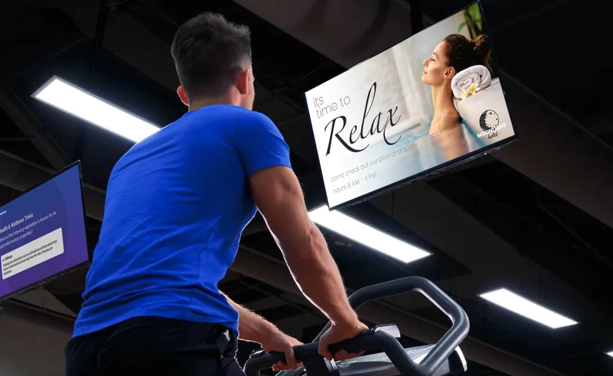 A man wearing a blue shirt uses a gym machine while looking up at a ceiling screen displaying an ad of a woman relaxing with towels and the text Its time to Relax. Fluorescent lights are visible overhead.