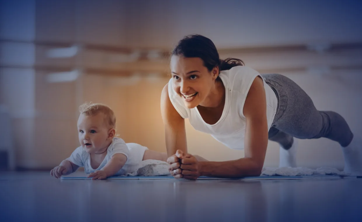 A smiling woman in workout clothes does a plank on a yoga mat beside a baby lying on their stomach in a bright, spacious room.